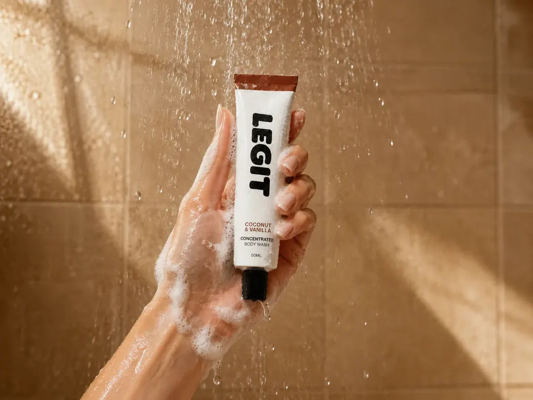 A hand holding a tube of Legit Concentrated Body Wash under the shower, covered in lather